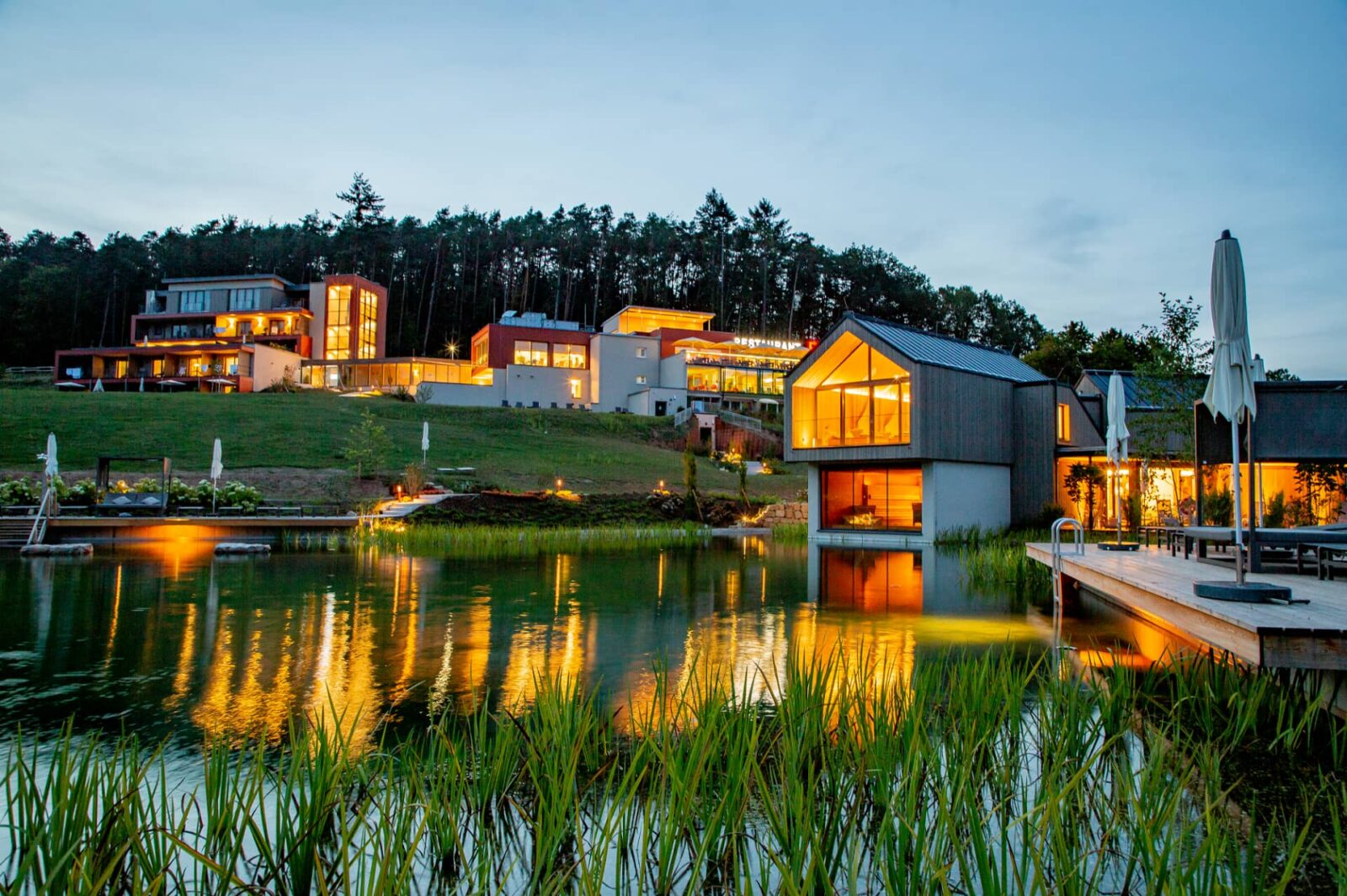 Außenansicht des beleuchteten Pfalzblick Wald Spa Resorts im Pfälzerwald mit Naturbadeteich und Sauna-Insel